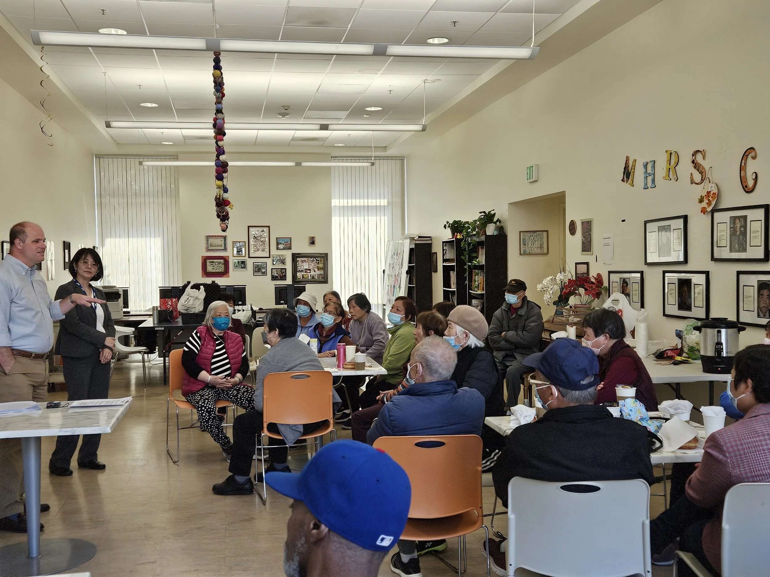 A group of seniors sit at tables in a classroom, while two people stand speaking at the front.