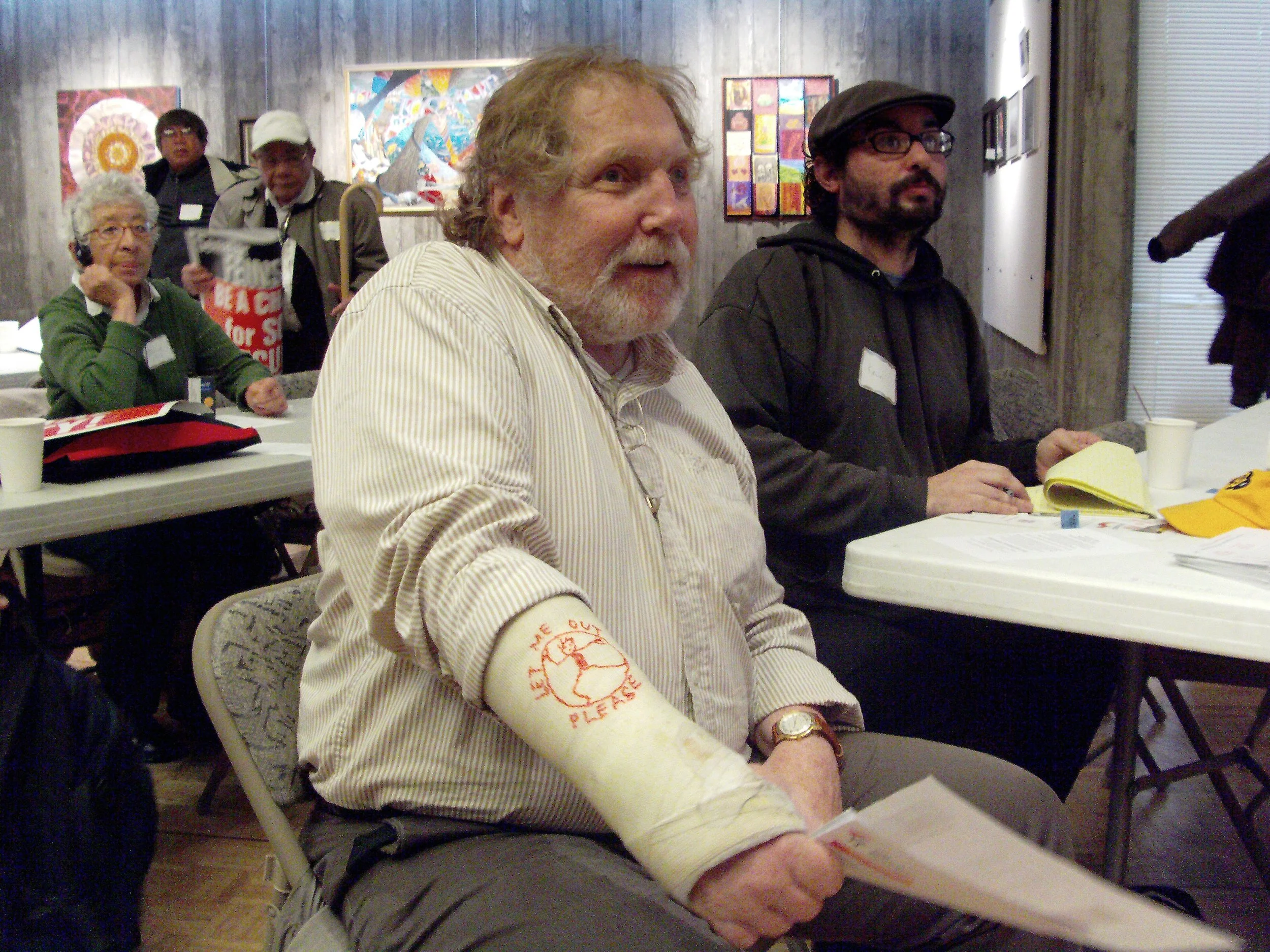 A man sitting at a table, smiling, with other people next to him and behind him. He is wearing a cast on his arm that says “let me out please"