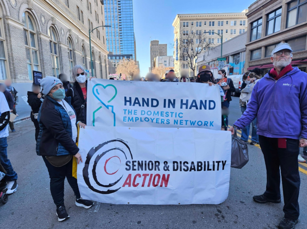 A group of people stands outside holding two banners, one that says Hand in Hand the Domestic Employers Network, and one that says Senior and Disability Action.