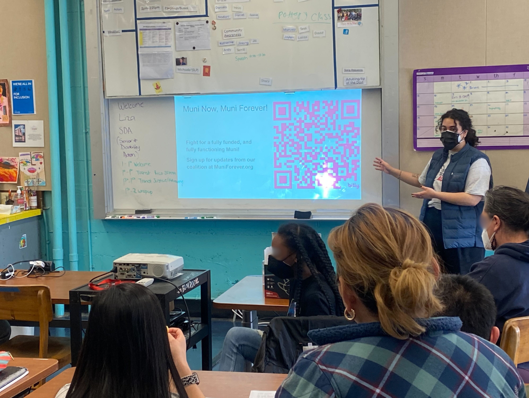 Liza stands at the front of a classroom and points to a whiteboard, with a powerpoint slide reading: “Muni Now, Muni Forever!”. A group of students, seated, face the whiteboard.