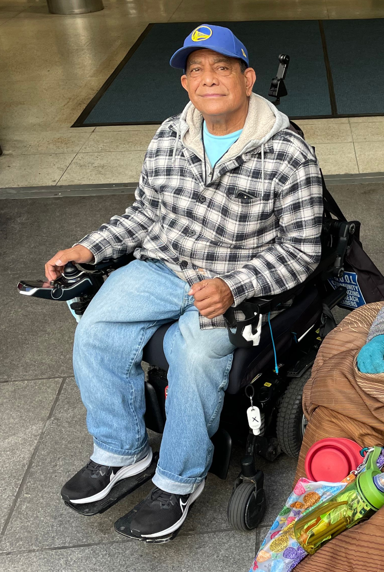 A man in a baseball cap sitting in a power wheelchair, smiling slightly