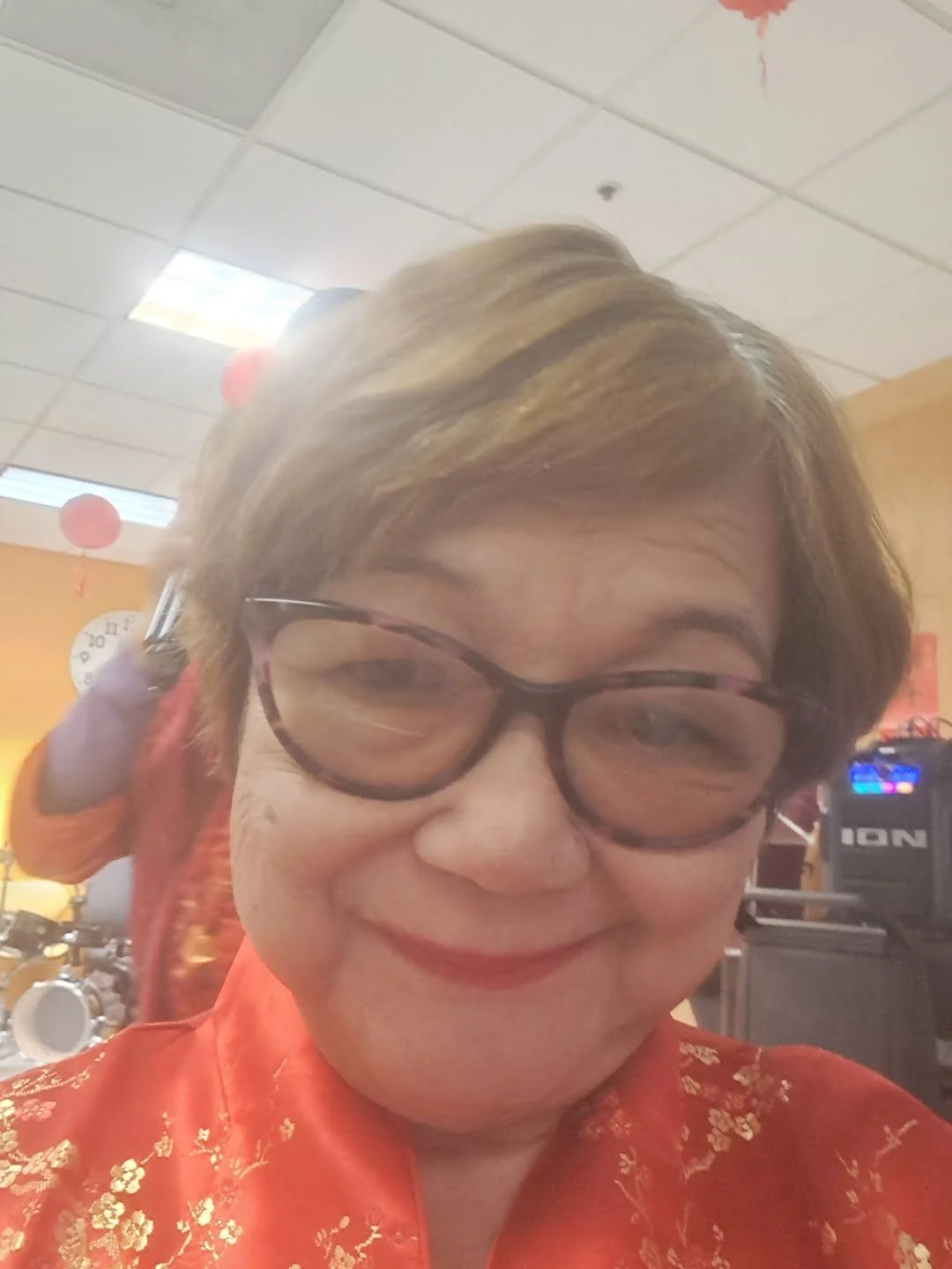 A woman smiling at the camera, with brown hair, glasses, and wearing an orange shirt with gold flowers