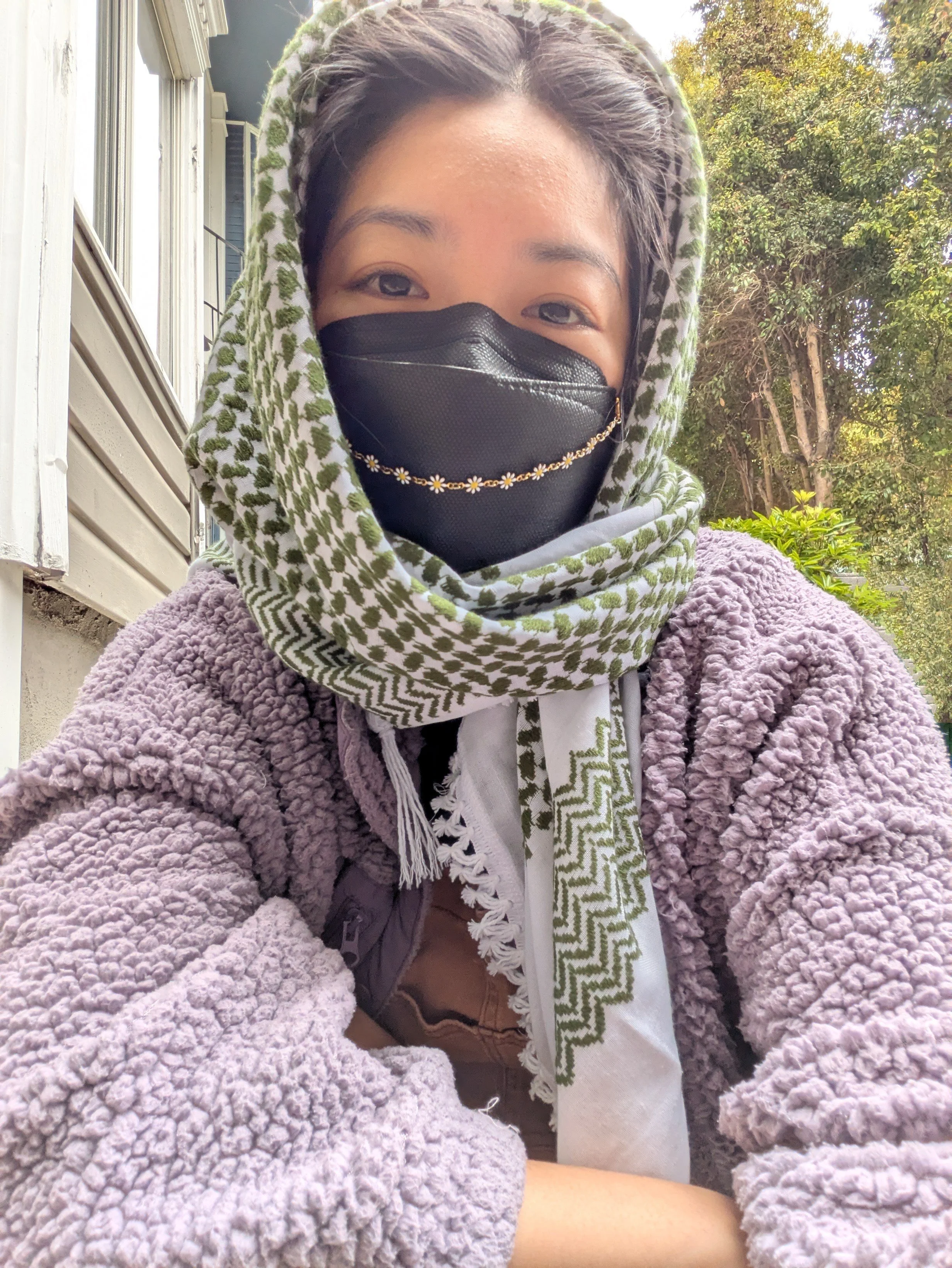 A person with black hair wearing a purple fluffy jacket, a keffiyah over their head, and a mask with jewelry on it.