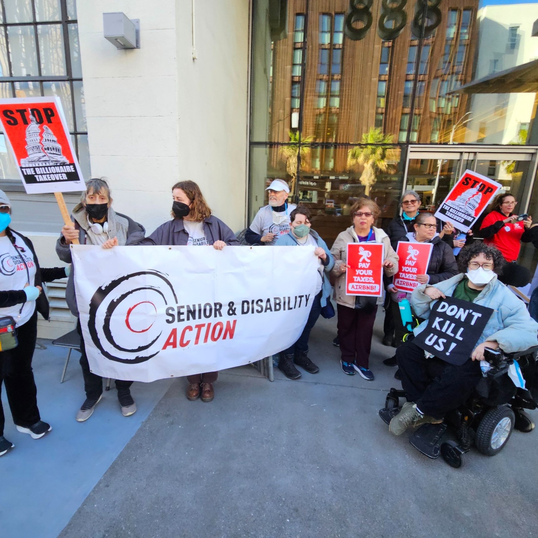 A group of masked seniors and people with disabilities holding signs that say “Stop the billionaire takeover” and “pay your taxes, Airbnb” in front of the SDA banner.