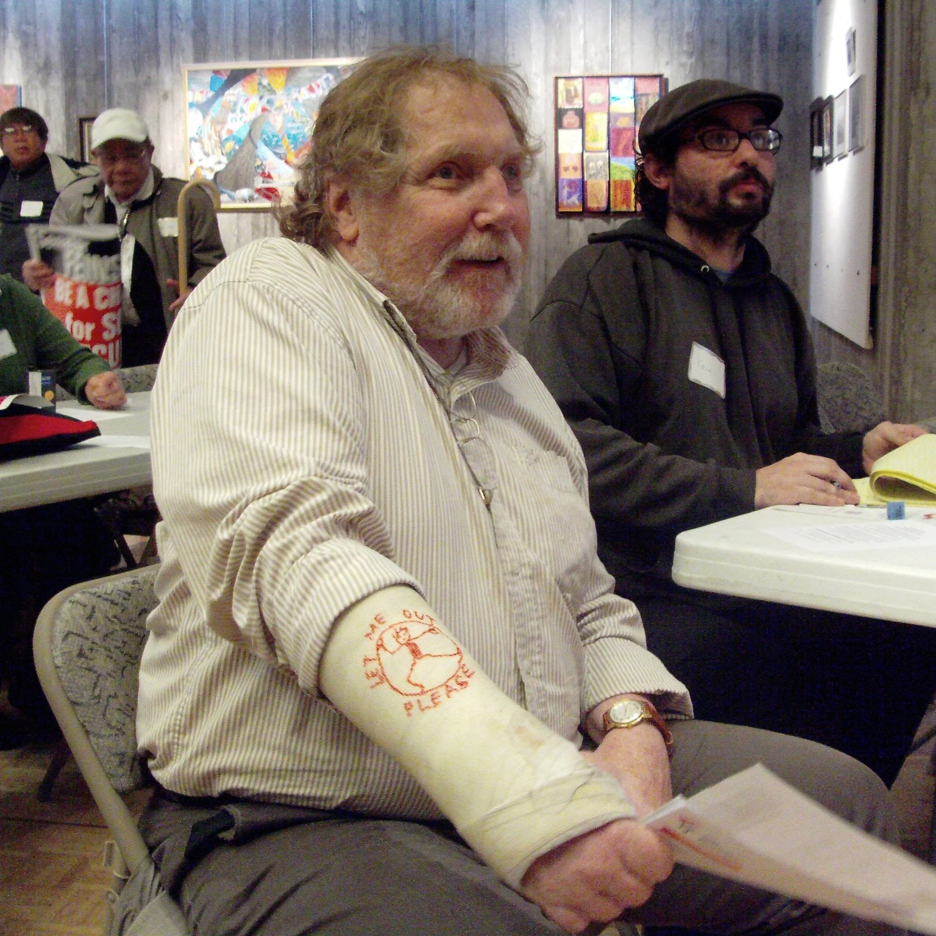 A man sitting smiling at a table with a cast on his arm that says "let me out please"