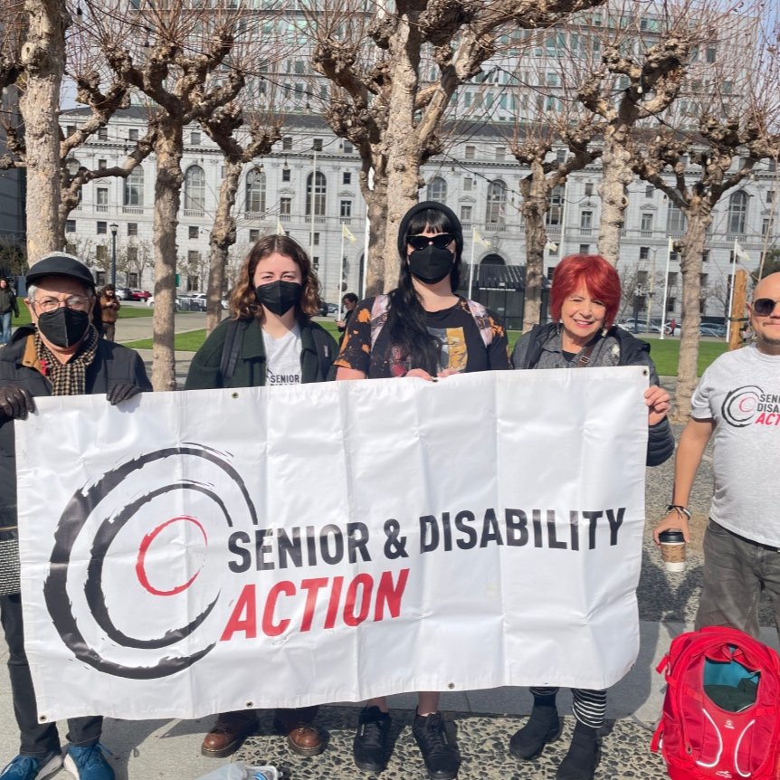 A group of five people, three of them wearing masks, stands outside in front of trees and holding a banner that says Senior and Disability Action