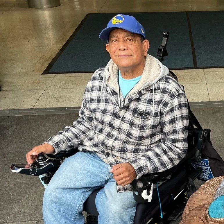 A man in a baseball cap in a power wheelchair, smiling slightly
