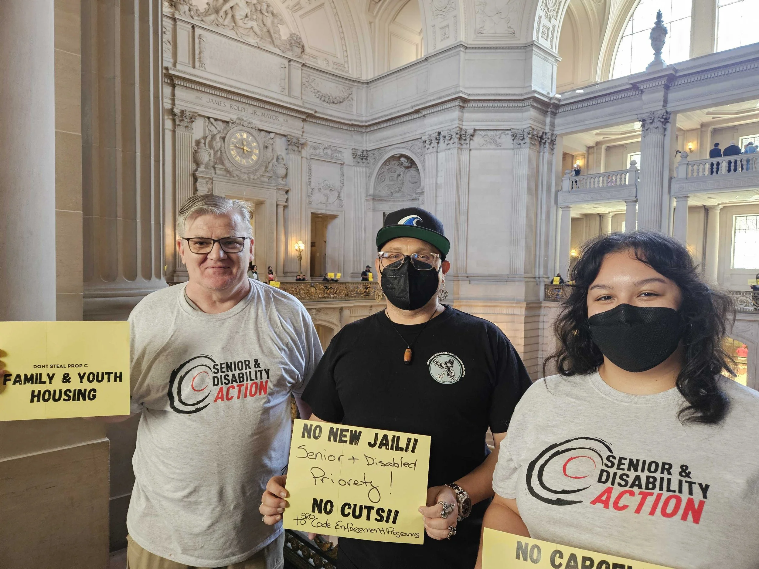 Three people standing in City Hall, two of them wearing SDA t-shirts. They are holding signs that say "No new jail! Senior & disabled priority! No cuts to SRO code enforcement programs." and "Don't steal prop C: family & youth housing"
