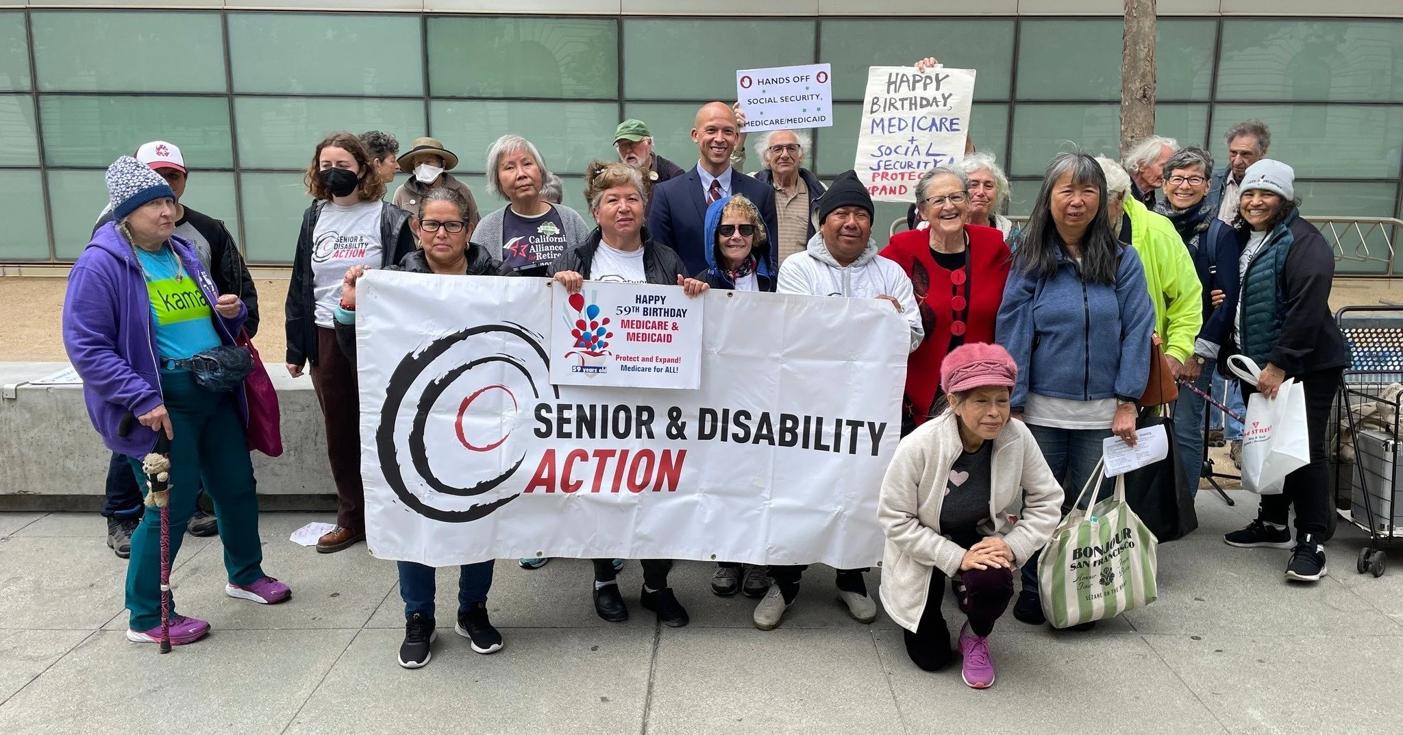 A group of people stands in front of a banner that says Senior and Disability Action. Others hold smaller signs that say Happy 59th Birthday Medicare and Medicaid and Happy Birthday Medicare & Social Security.