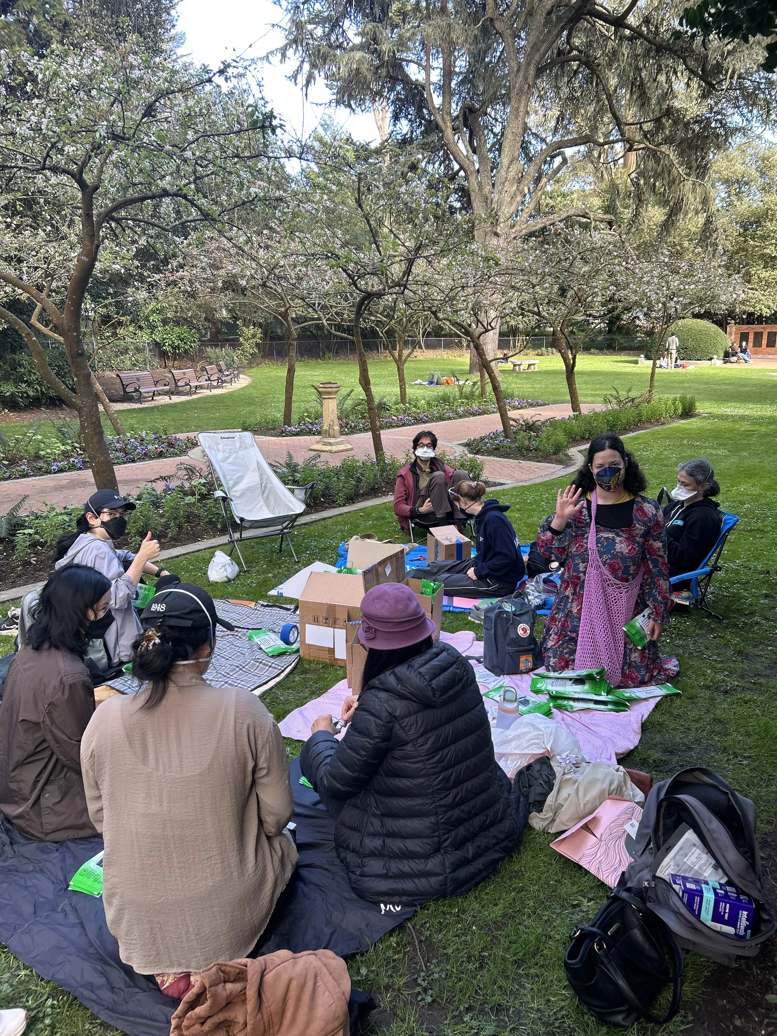 Un grupo de personas enmascaradas sentadas afuera en un parque, con cajas de máscaras a su alrededor.
