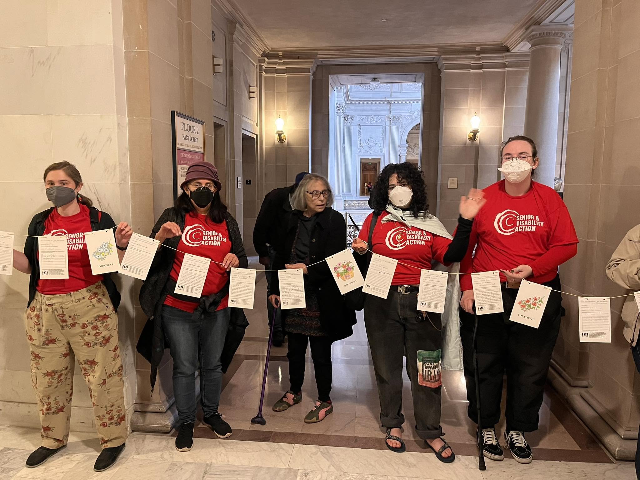 Un grupo de cinco personas, la mayoría enmascaradas y con camisetas de SDA, sosteniendo una serie de postales (no legibles en la foto). Están en la Alcaldía, con paredes de piedra gris detrás.
