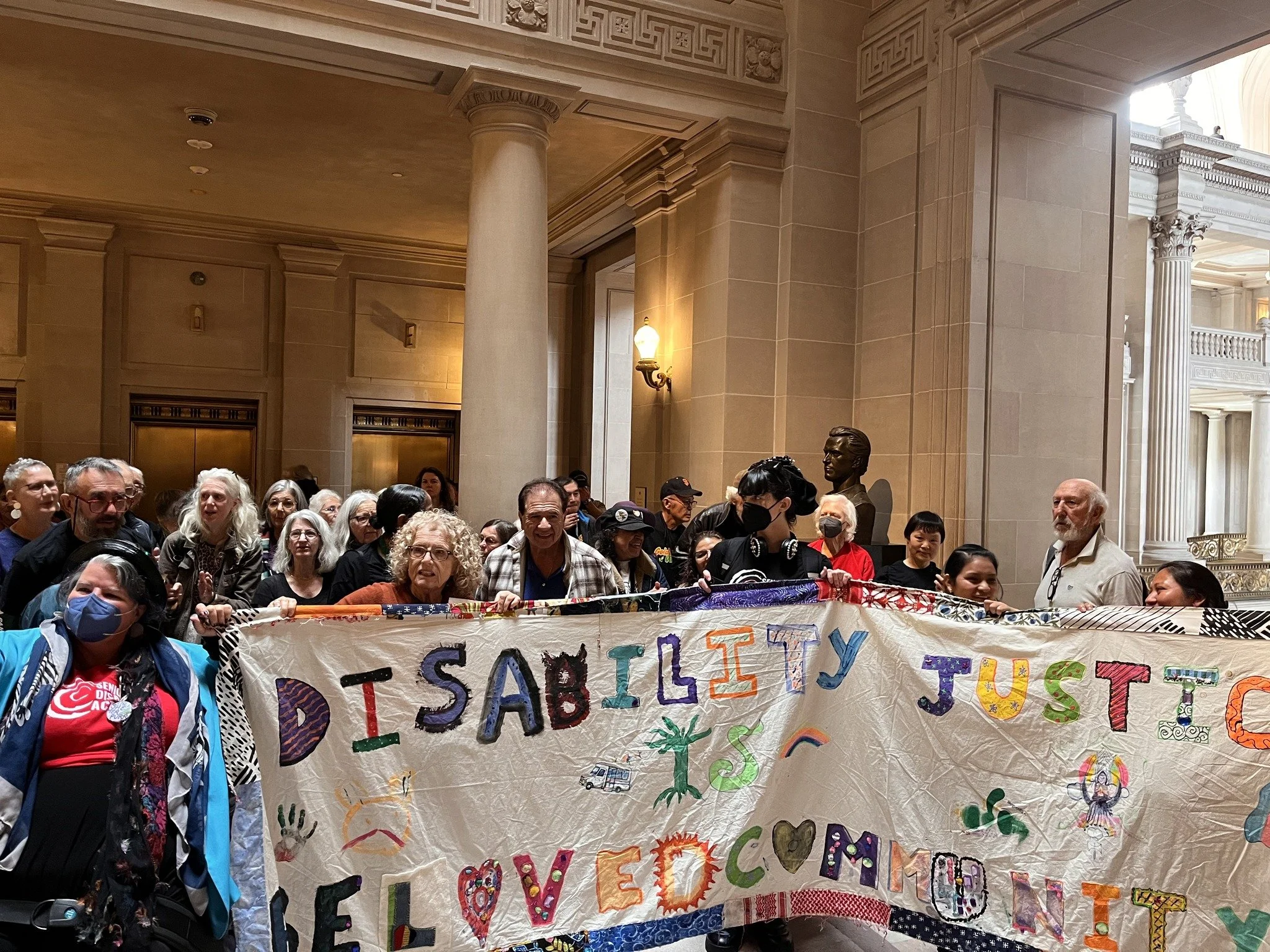 A large group of people in City Hall behind a fabric banner that says "Disability Justice is Beloved Community"