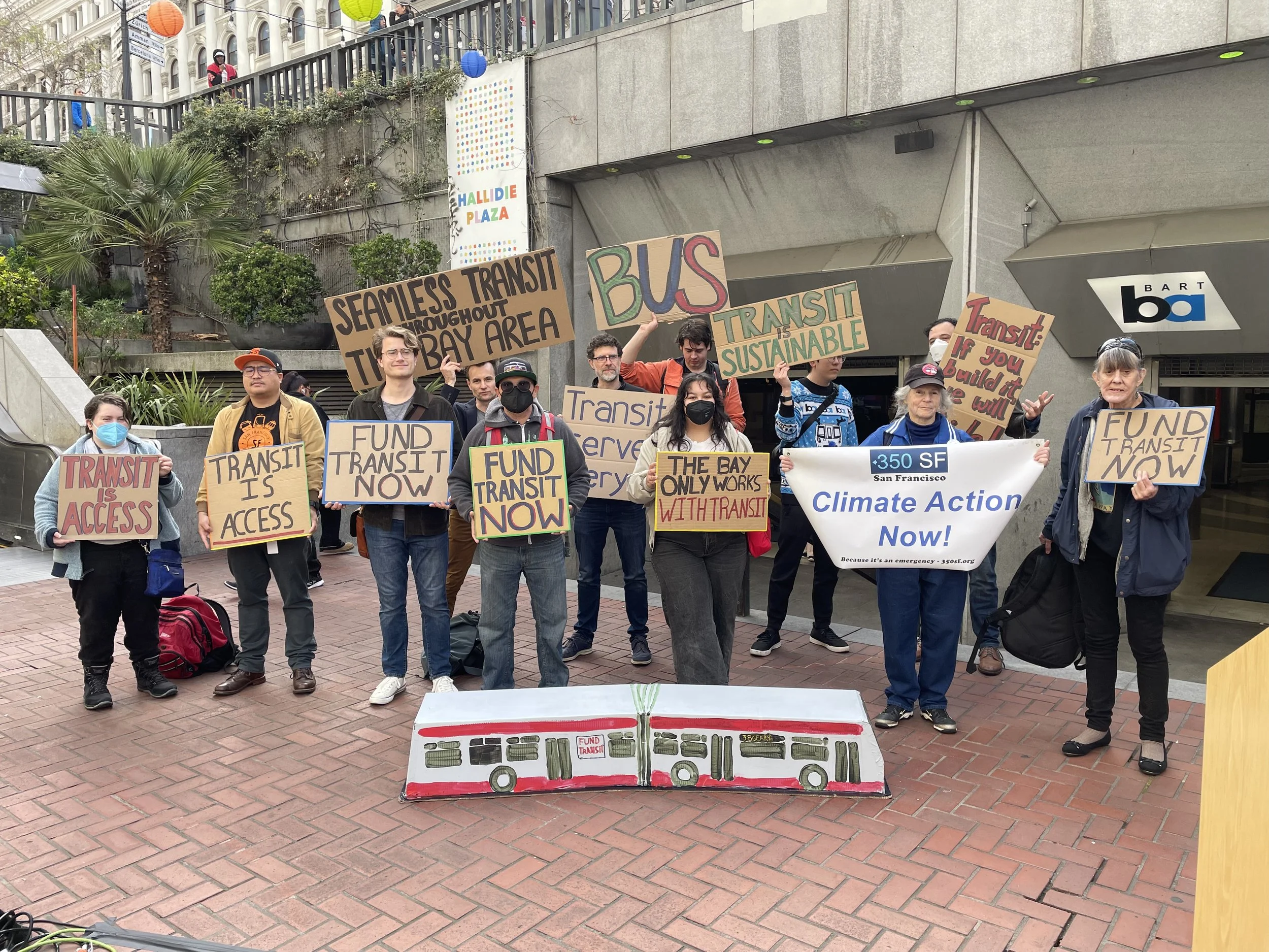 A group of around 10 people holding signs that say things like “transit is access” and “fund transit now.” There is a cardboard model of a MUNI bus on the ground in front of them.