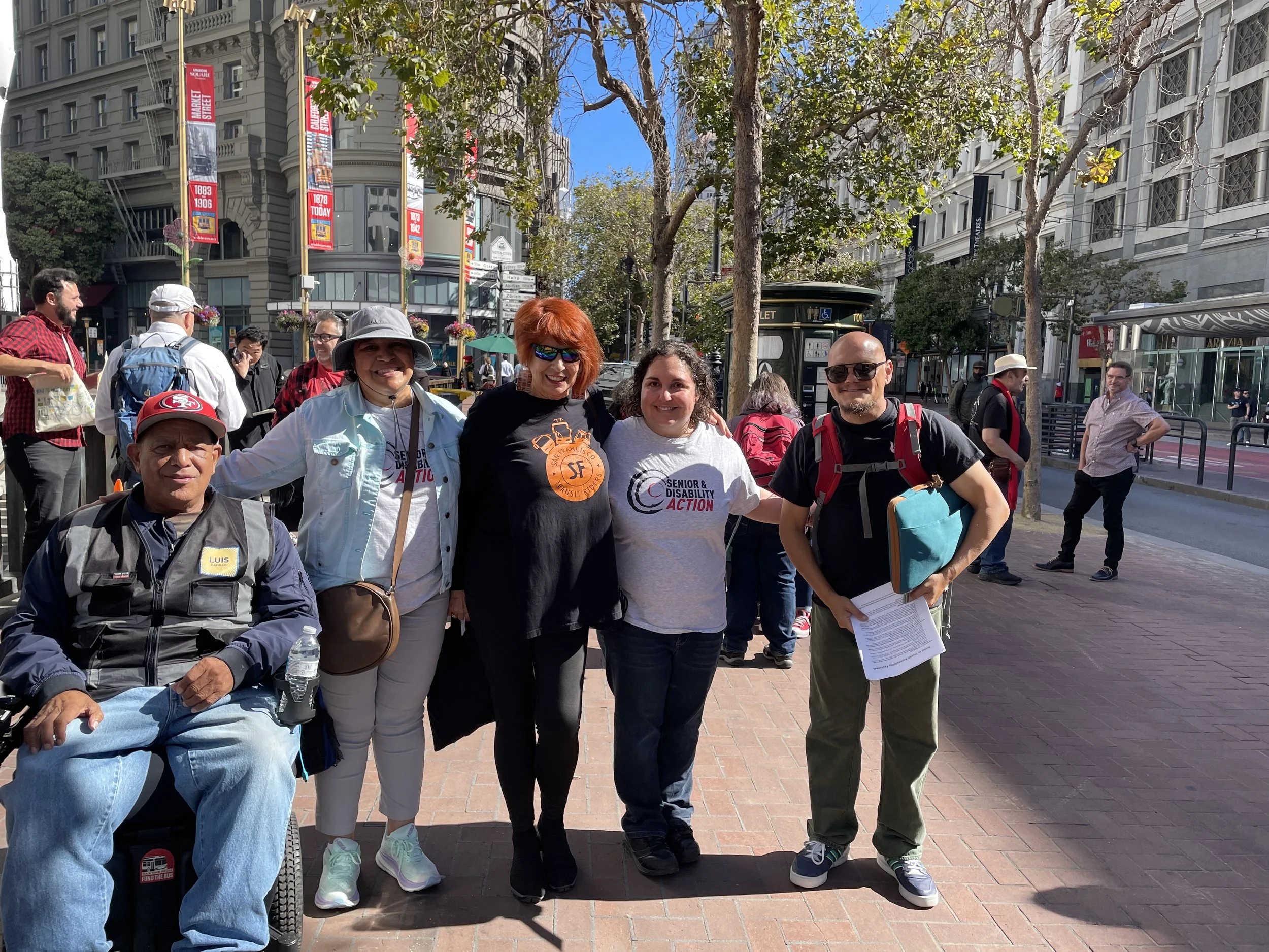A group of five people, one of them in a wheelchair and four standing on Market Street, wearing signs for SDA and SF Transit Riders