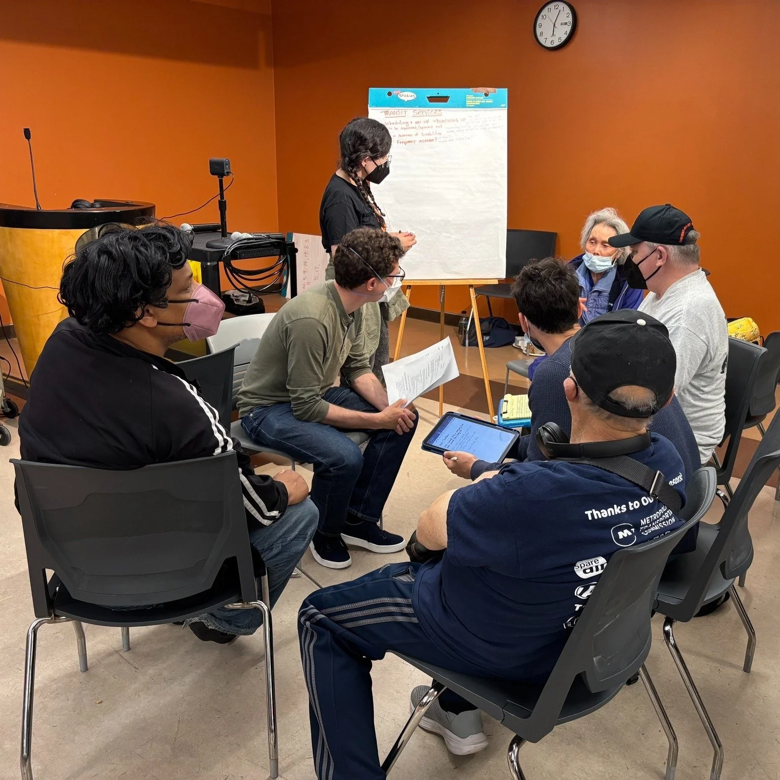 A small group of seven people, all wearing masks, sits in conversation, as one person stands at an easel taking notes on a large sheet of paper.