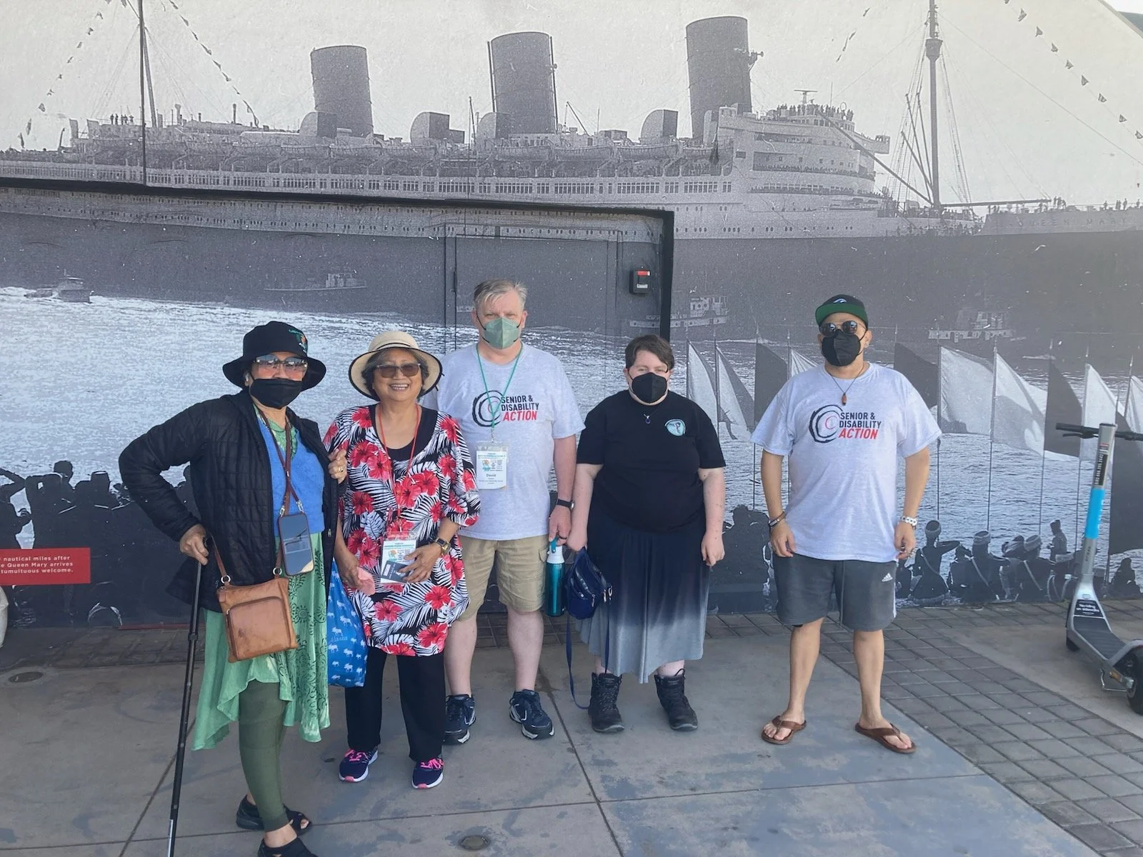 A group of five people, three of them masked and two of them in SDA shirts, standing in front of a large picture of a boat
