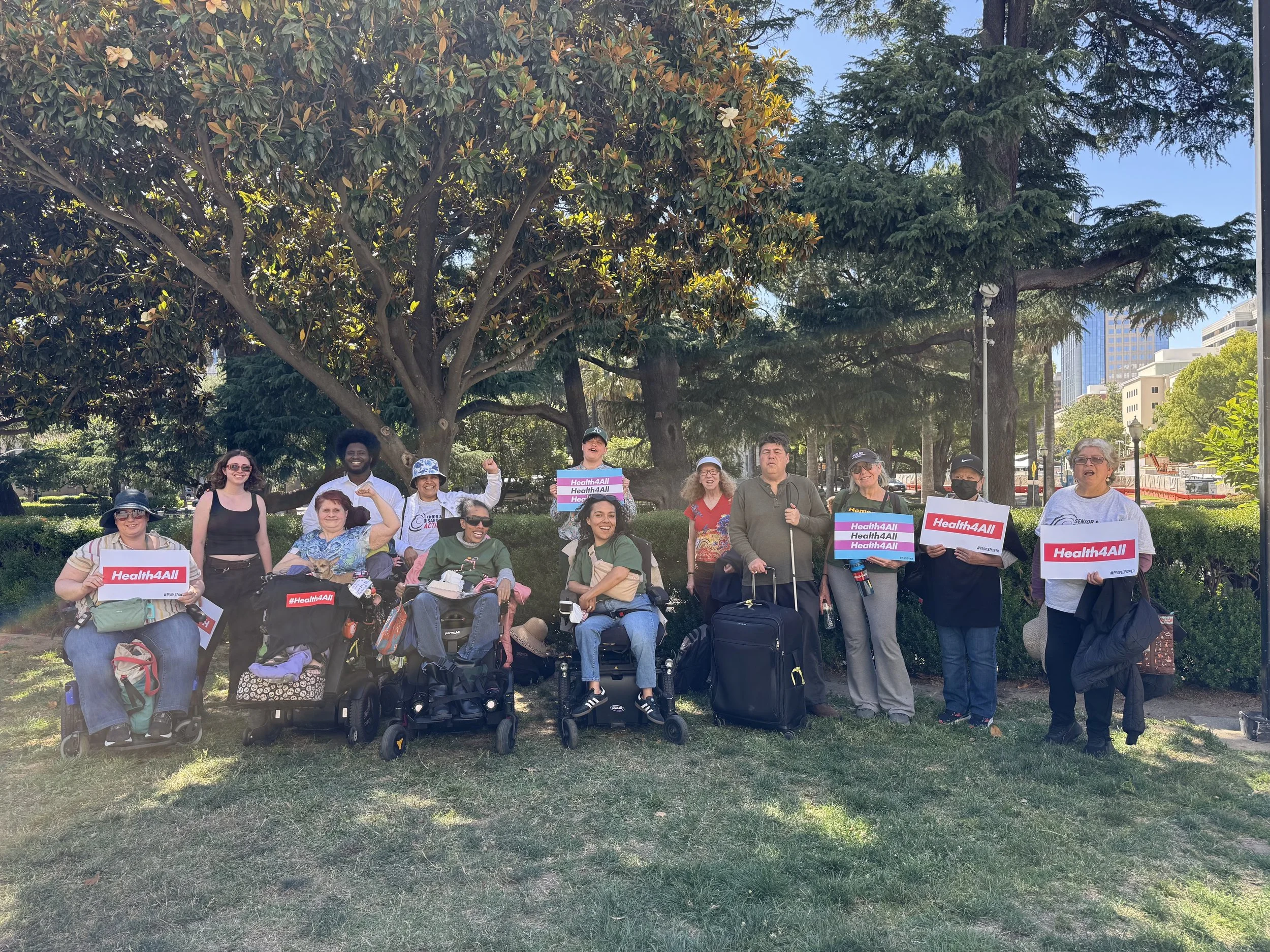 A group of seniors and disabled people, some standing and some in wheelchairs, outside on a lawn holding signs that say “Health4All”