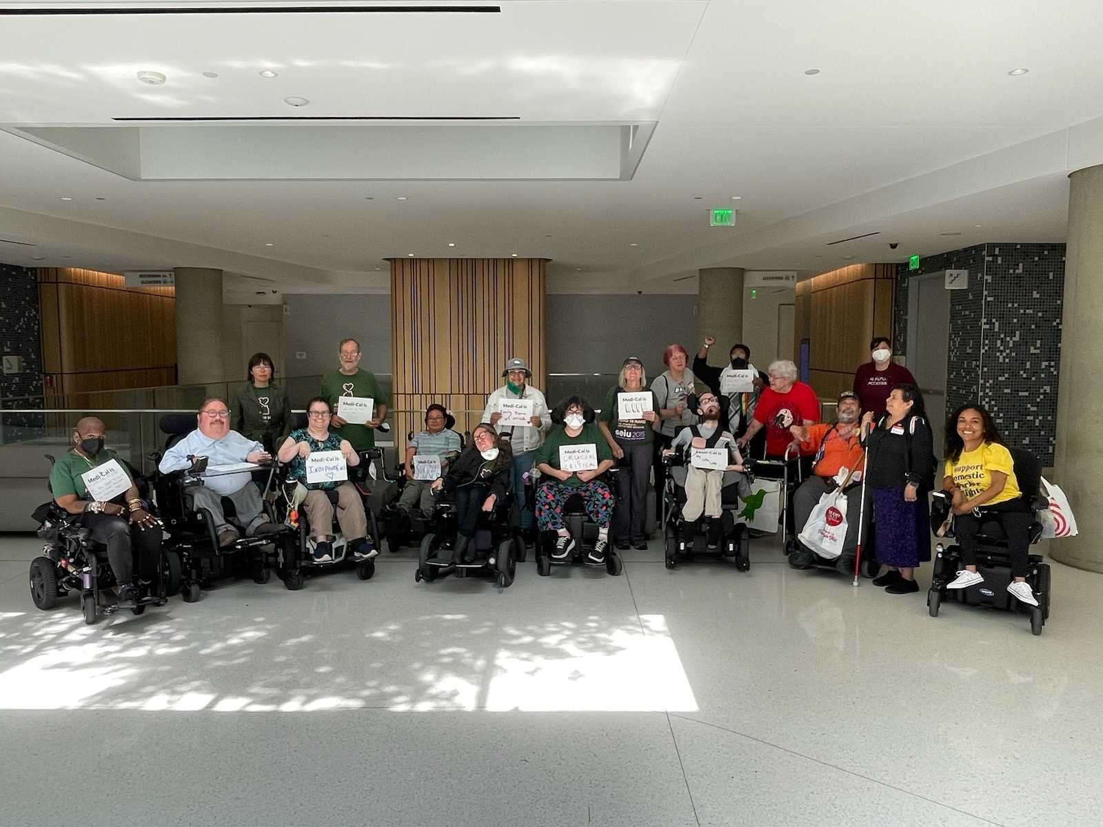 A group of people standing, in wheelchairs, and using other mobility devices, holding signs that say “Medi-Cal is…” and the most visible sign says “Medi-Cal is crucial and critical"
