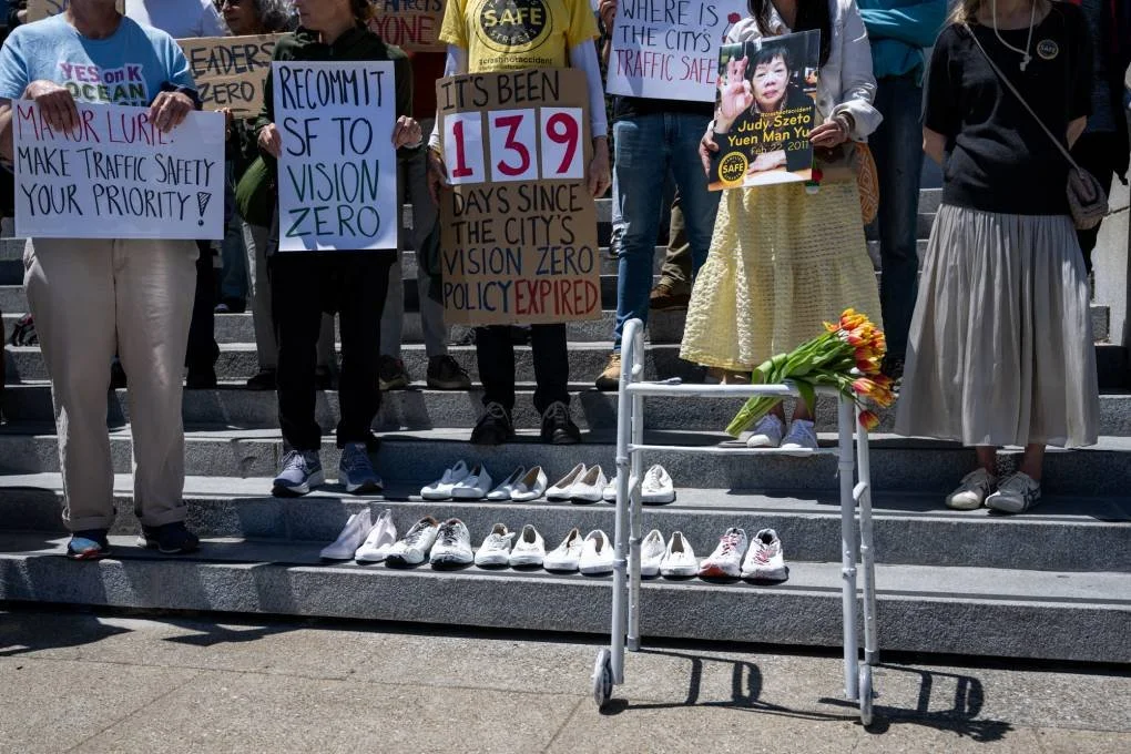 People standing on steps, holding signs that say “Recommit SF to Vision Zero” and “Mayor Lurie, make traffic safety your priority”. On the steps in front of them are ten empty pairs of shoes and a walker with flowers on it.