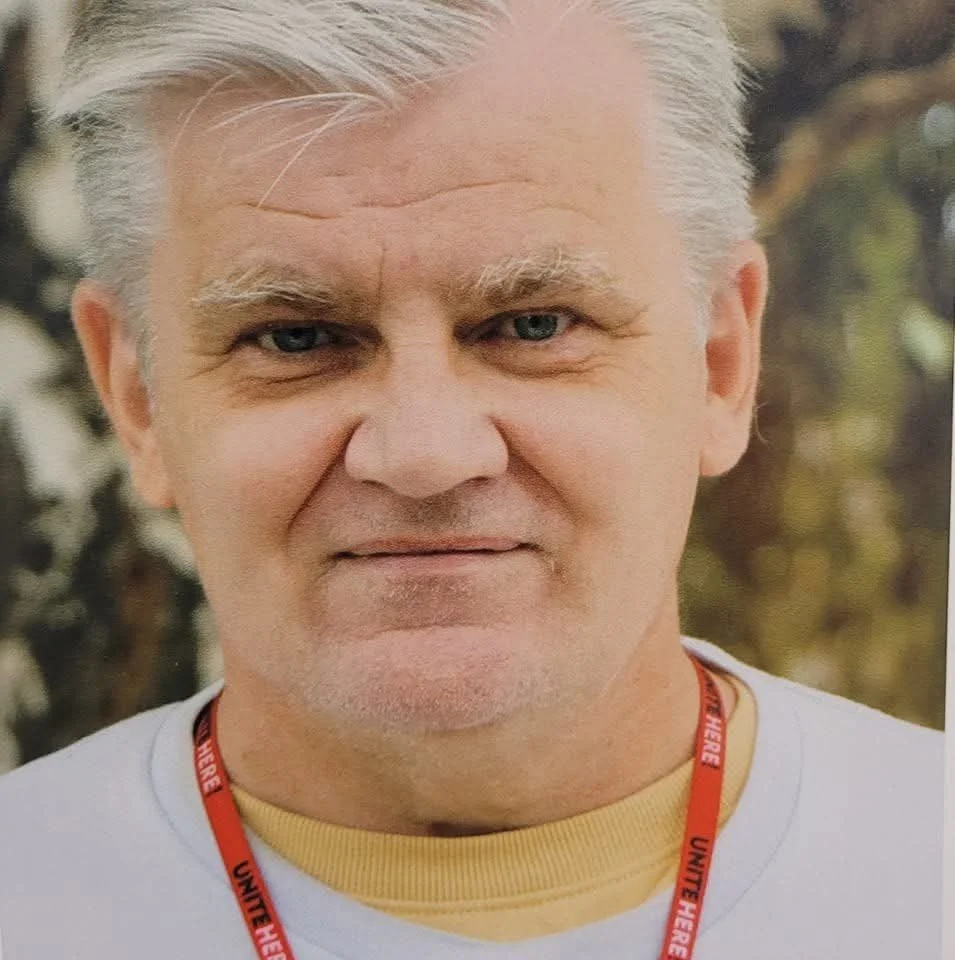 A person smiling slightly with white hair and a necklace that says Unite Here