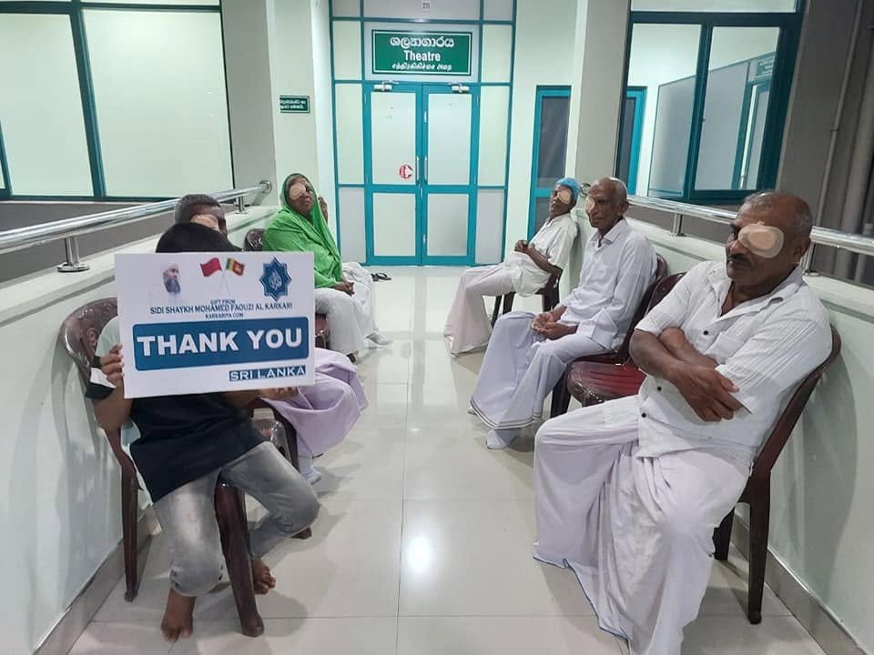 Group of four elderly men and two women sitting in a hospital corridor, with one child holding a 'Thank You Sri Lanka' sign in front of the group.