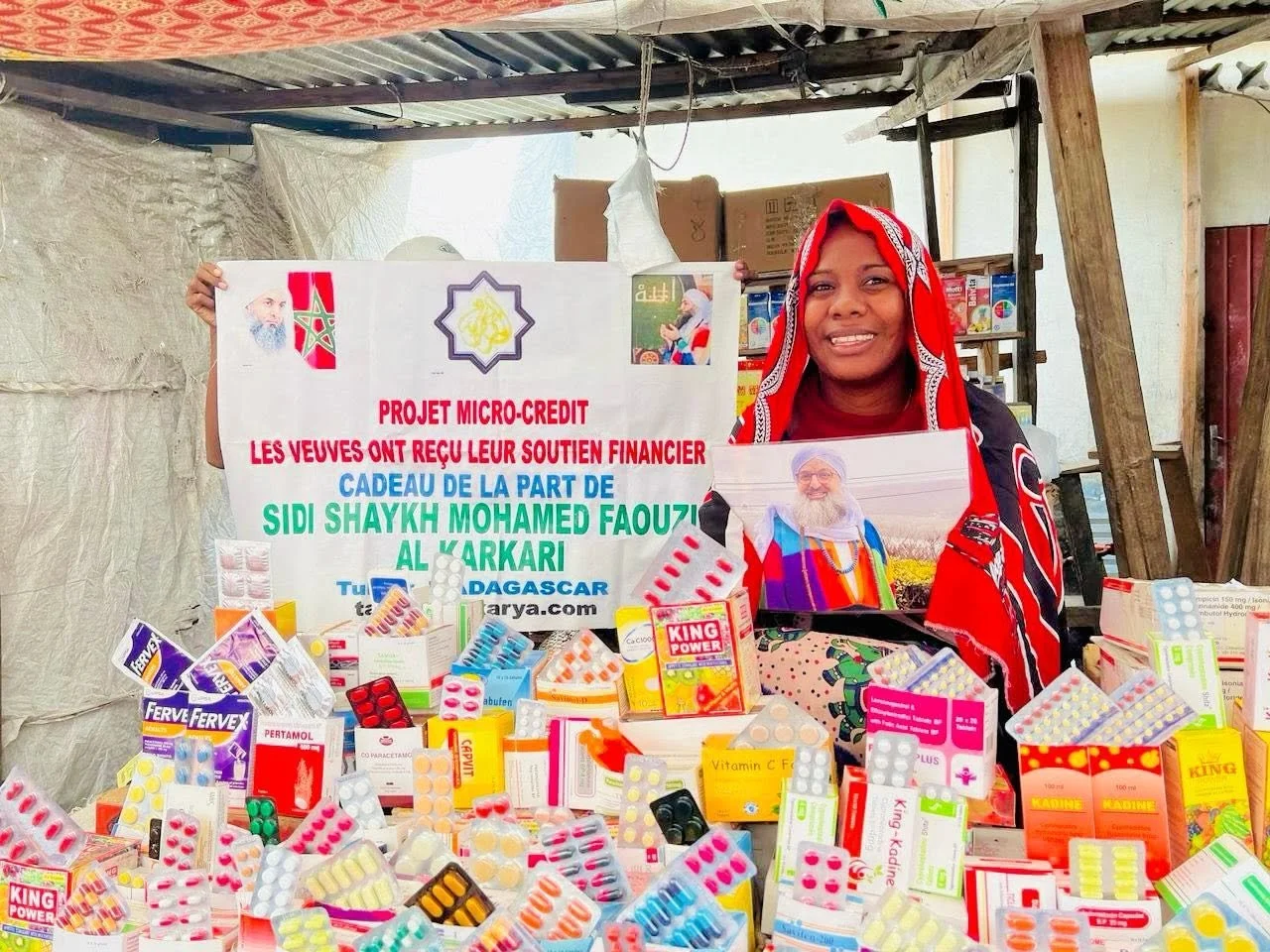 A woman smiling at a community health stall with various medicine packages, holding a sign in French about a micro-credit project and a photo of Sheikh Mohamed Faouz.