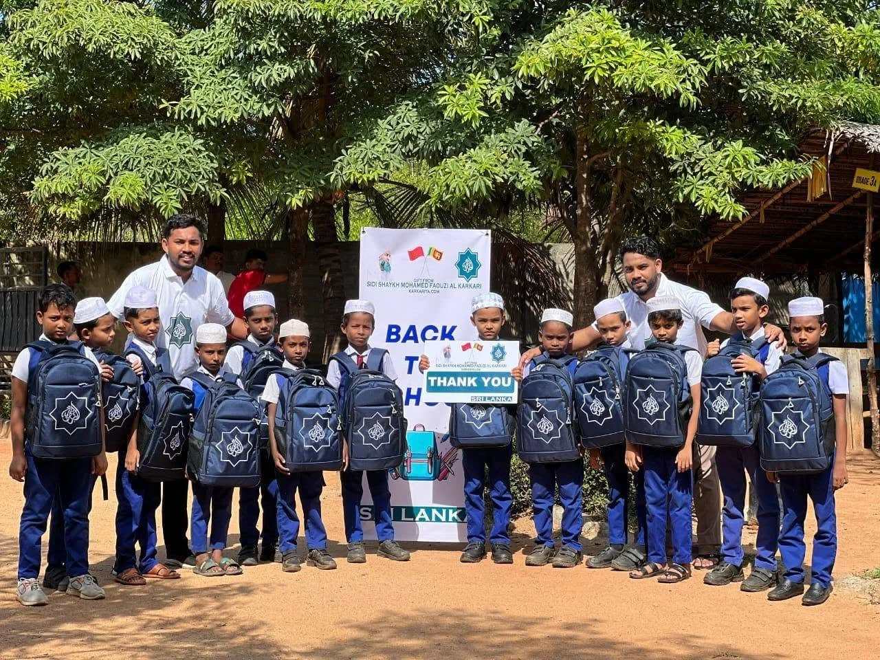Group of young boys in school uniforms holding new backpacks, standing with two adults outdoors near trees, in front of a sign that says "Back to School" and "Thank You".