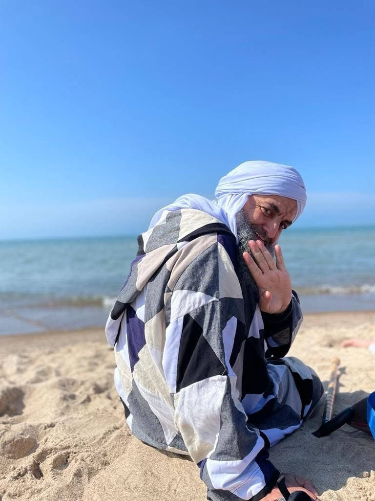 A man sitting on a sandy beach near the ocean, wearing a white headscarf and a colorful, geometric-patterned jacket, with his hand held up to his face.