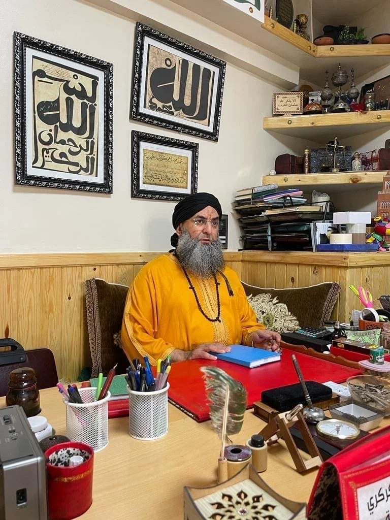 A man wearing a black turban and yellow traditional garment sitting at a cluttered desk in a room decorated with framed Arabic calligraphy on the wall.