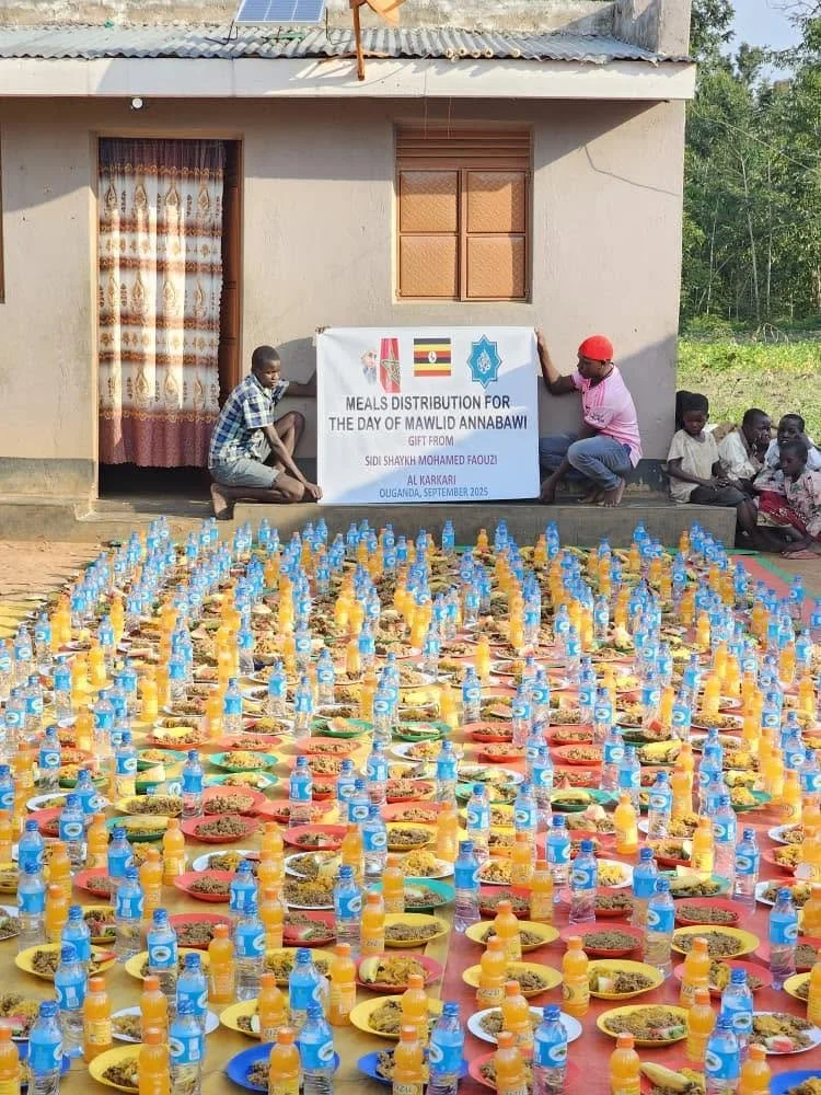 Outdoor scene with children and adults preparing a large distribution of meals and beverages in Africa, with plates of food and bottles of water and juice arranged in rows on the ground. A house with a curtain-covered doorway and a window is in the b