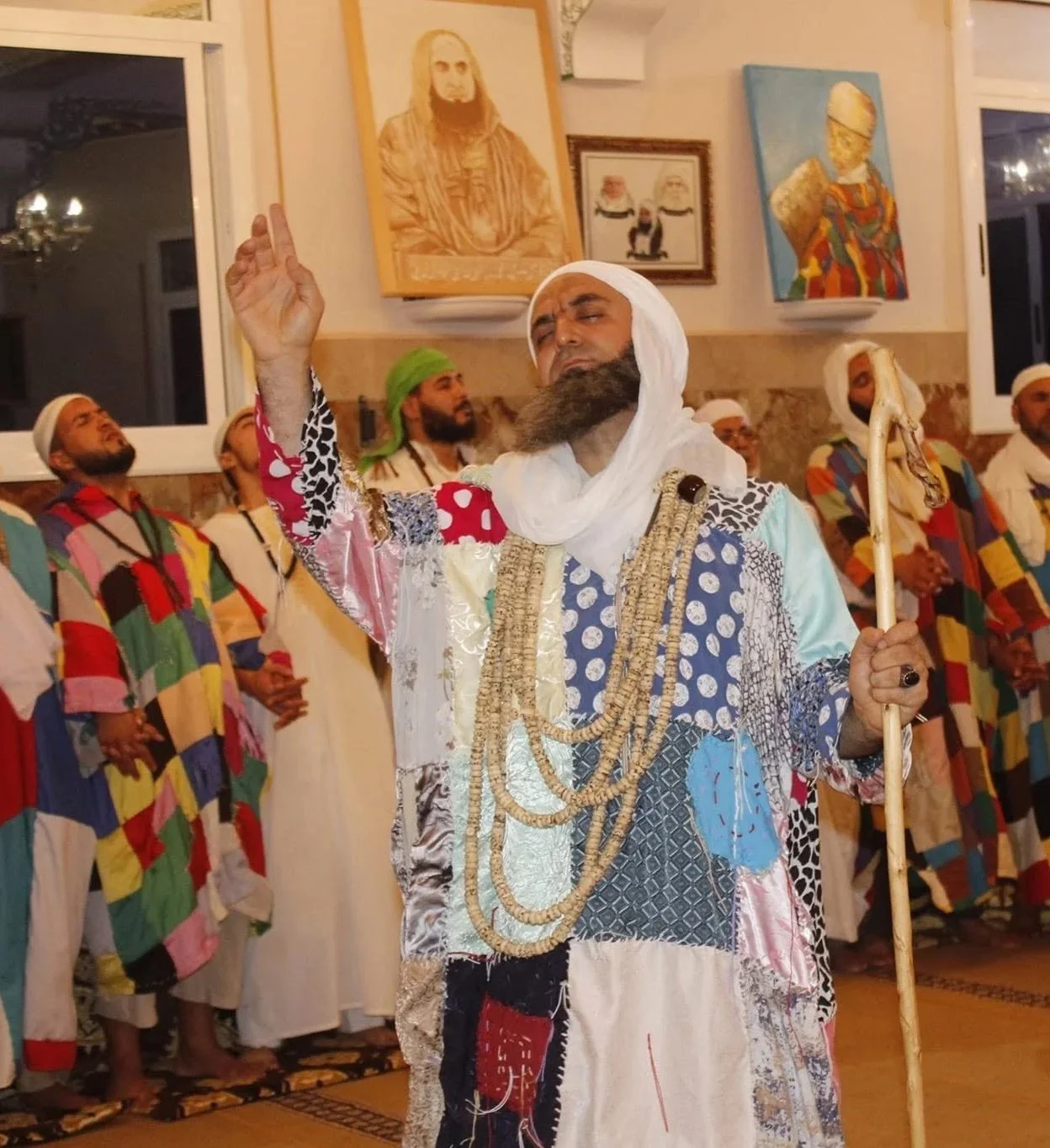 A man dressed in colorful patchwork clothing and adorned with multiple necklaces and a white headwrap, performing a traditional dance or ritual with his eyes closed, while holding a staff in his right hand. Behind him, several men in multicolored pat