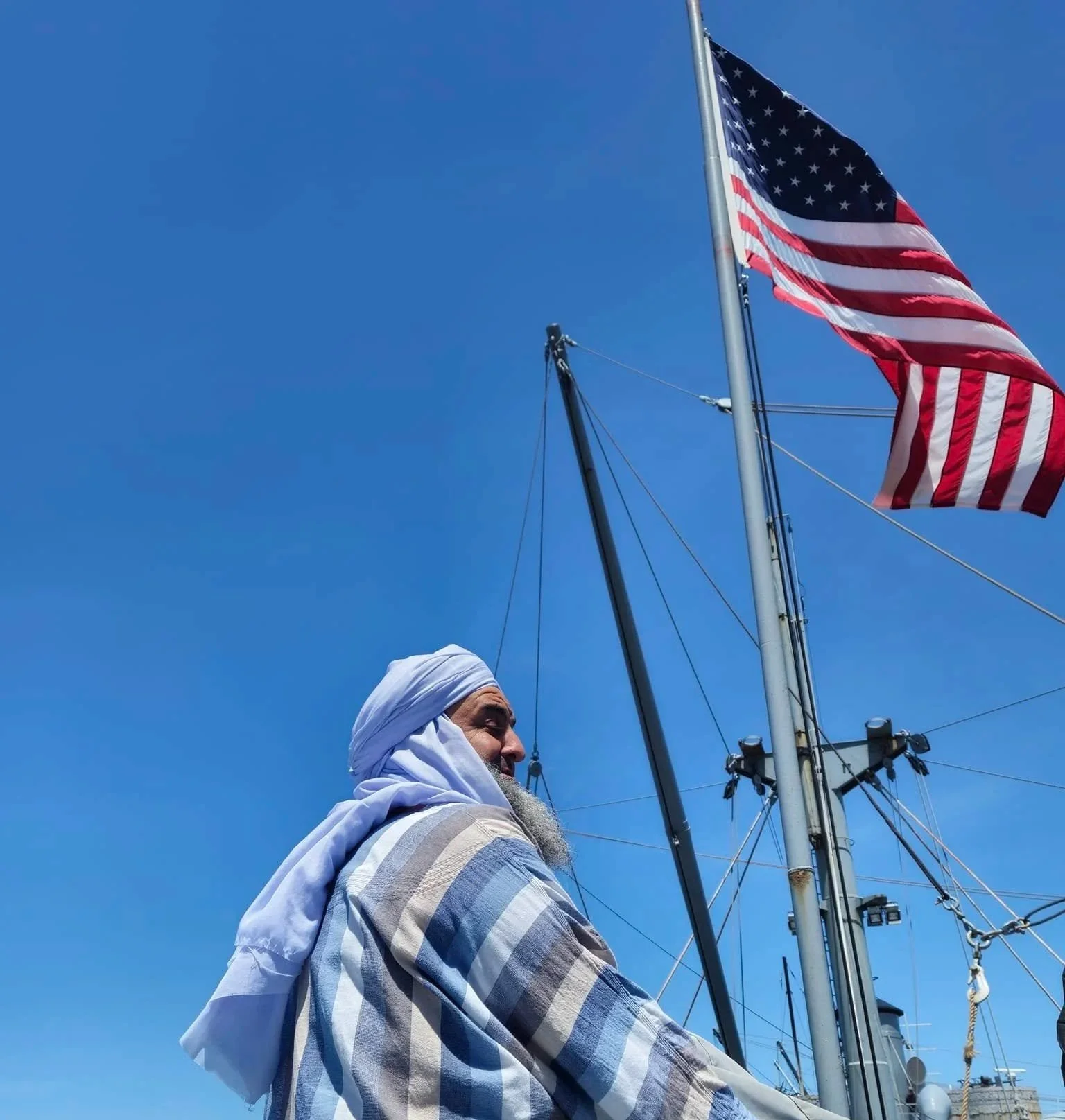 A man wearing a white head covering and a striped shirt standing on a boat, with the American flag flying on a tall flagpole against a clear blue sky.