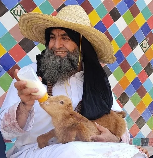 A man with a beard wearing a large straw hat, white clothing, and a black cloth over his head, holding a young brown goat and bottle-feeding it with milk. The background features a colorful, diamond-patterned wall.