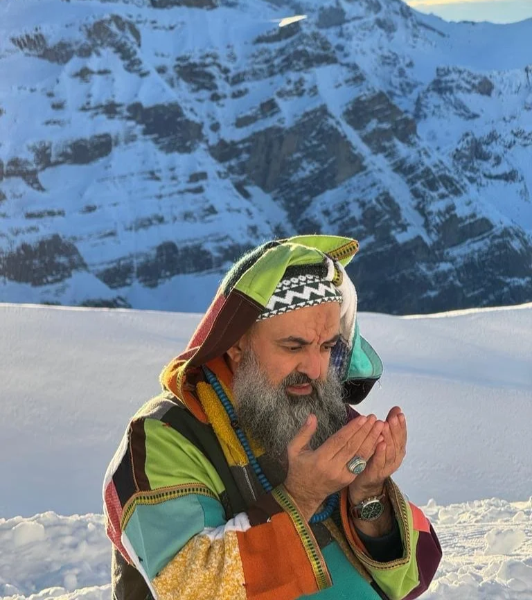 A man with a gray beard outdoors in a snowy mountain landscape, wearing a colorful patchwork jacket and a patterned headband, holding his hands close to his face.