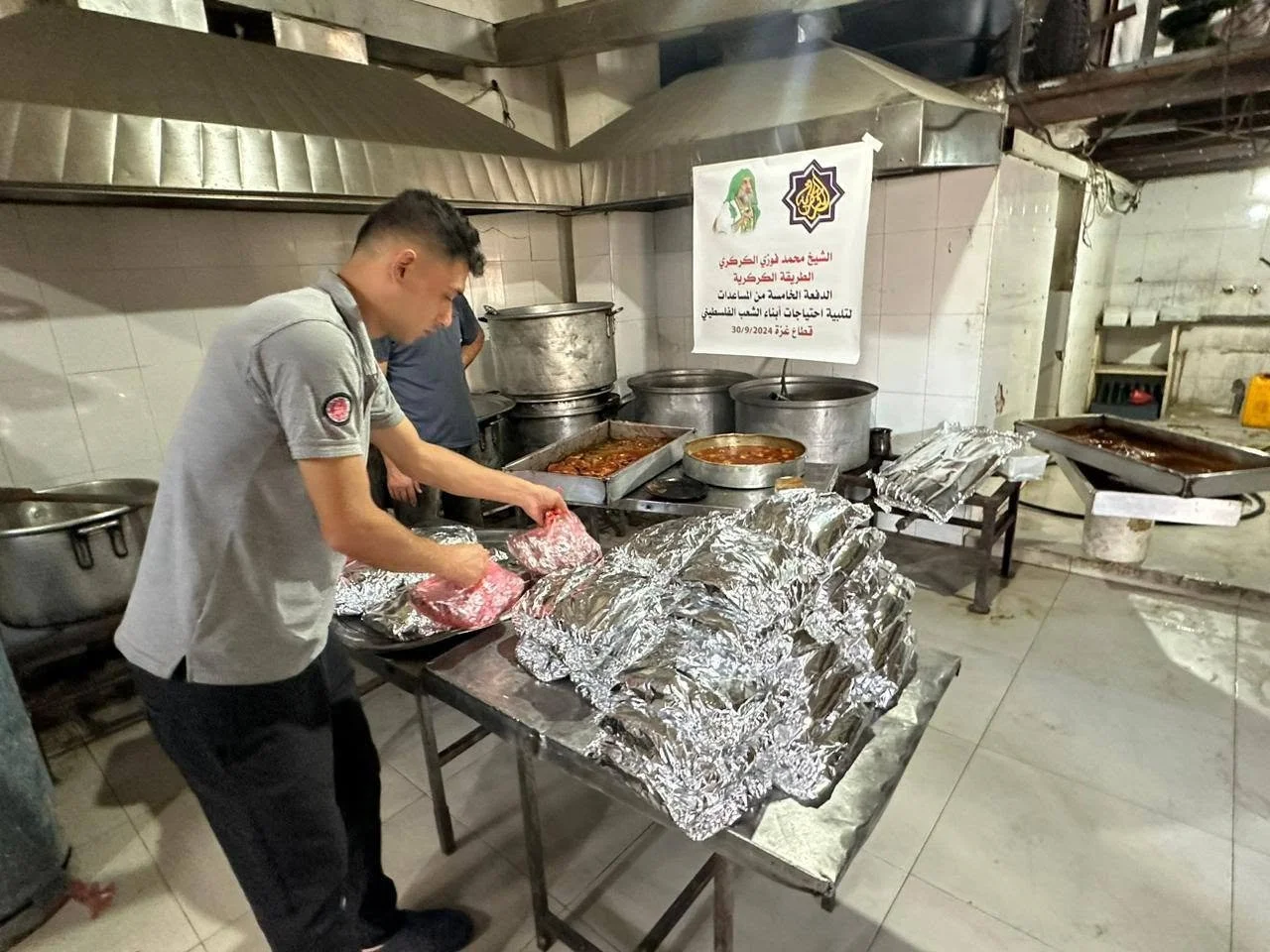 A man preparing food in a commercial kitchen with large pots and trays of cooked dishes. There are aluminum foil-wrapped food items on the table, and a banner with Arabic writing and logos on the wall.
