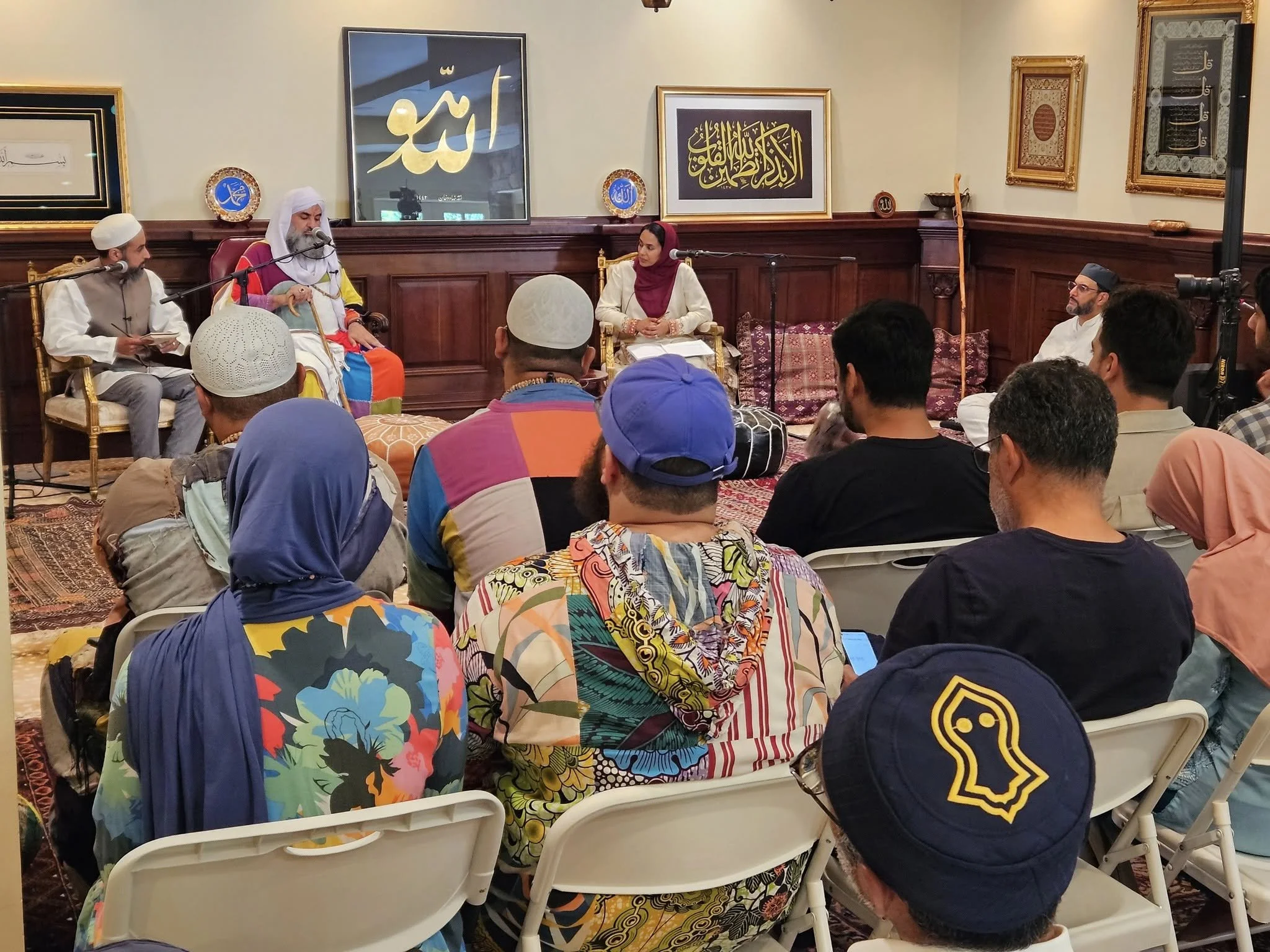 A group of people attending a religious or cultural event, seated in chairs, facing a panel of speakers in a room decorated with framed religious calligraphy.
