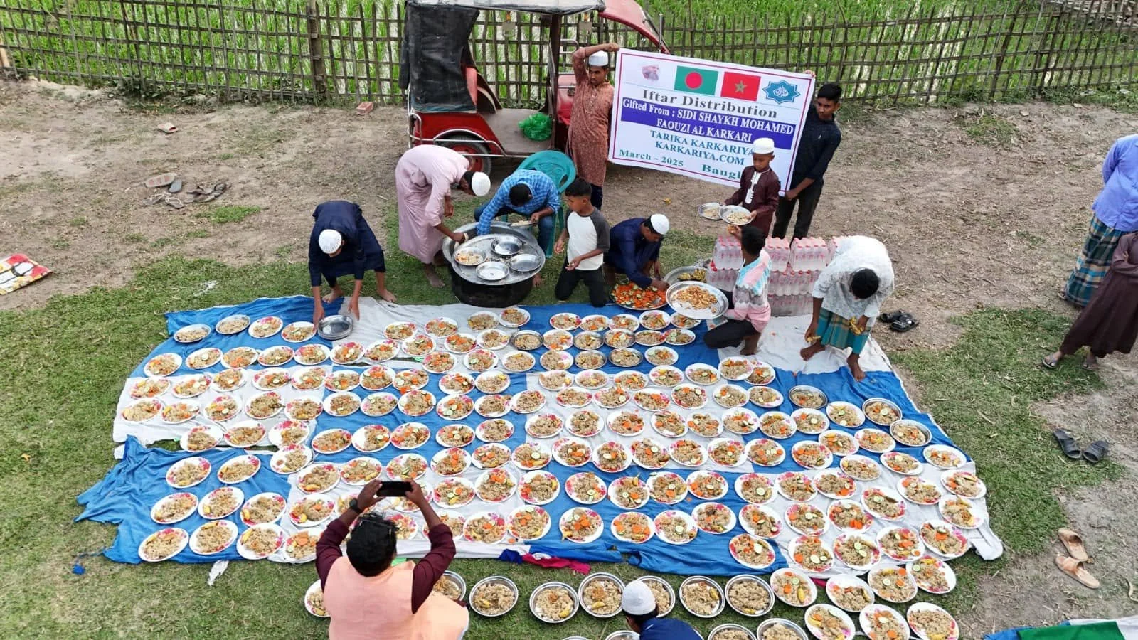 A large outdoor community meal distribution event with many plates of rice and food laid out on blue and white sheets, with several people serving and organizing the food, and a banner in the background indicating it is an Iftar distribution event.
