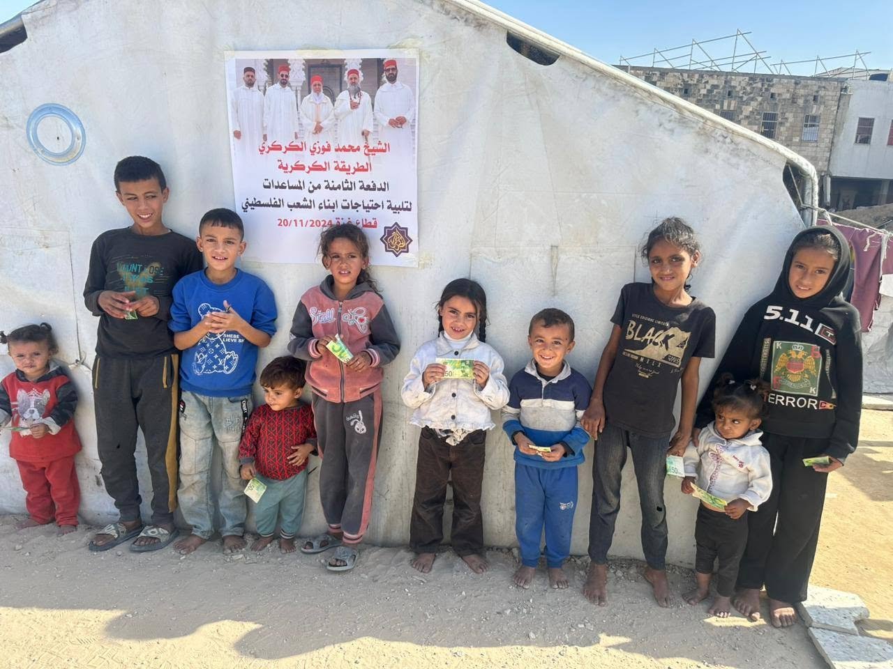 A group of ten children standing against a white wall, holding money and smiling. There is a poster with Arabic text above them on the wall.