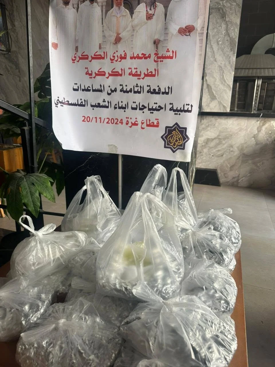 Plastic bags filled with food or supplies placed on a table, with a sign in Arabic above them indicating they are for supporting Palestinian children on 20/11/2024, organized by Sheikh Mohammed Fuzi Al-Kurky.