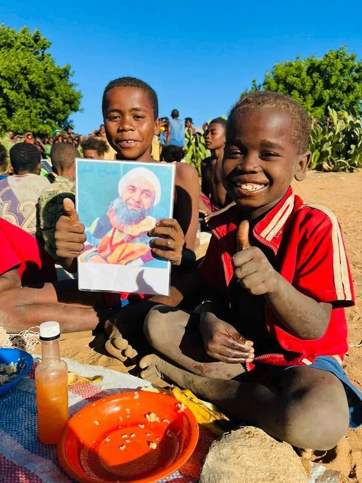 Two smiling boys sitting outdoors on the ground, one holding a picture of a bearded man in religious attire, giving a thumbs-up. There are other children in the background, some sitting on the ground, with trees and a clear blue sky overhead. On the 