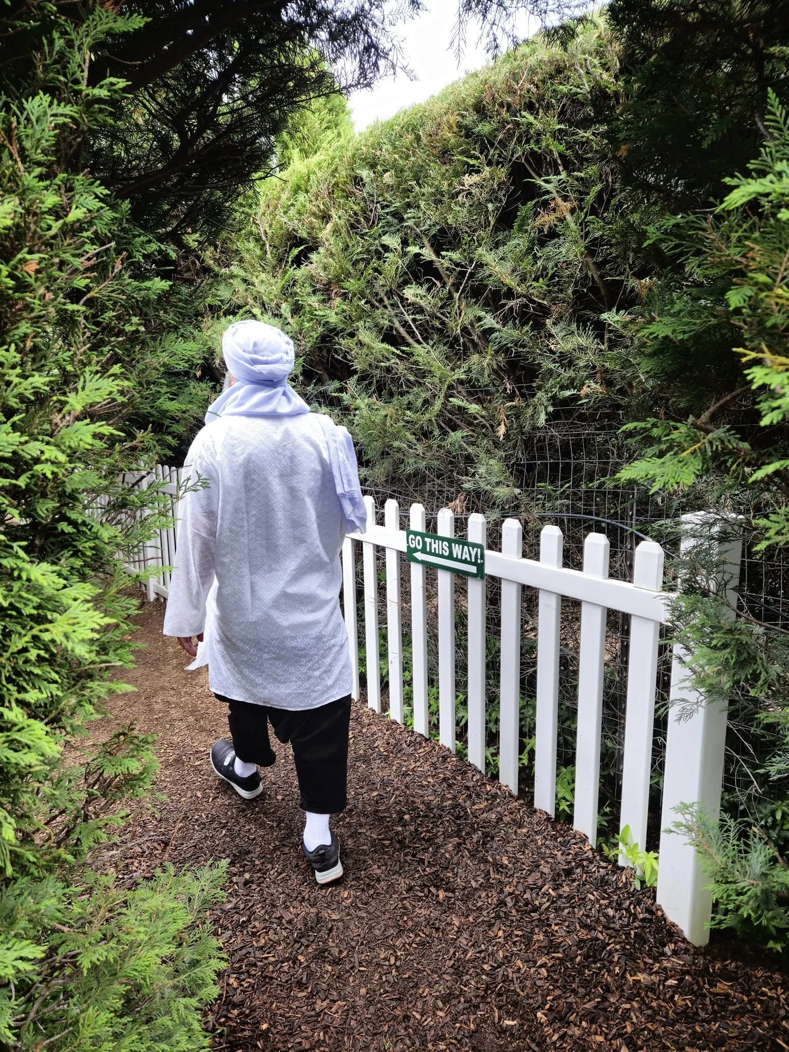 A person dressed in white with a headscarf, walking on a dirt path between greenery and a white fence with a sign that says 'Go This Way!'