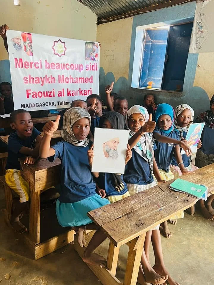 Group of smiling children in a classroom, holding a sign expressing thanks to Sheikh Mohamed Faouzi al Karkari and a portrait of a bearded man. The children are in blue uniforms with headscarves or hair visible, sitting at wooden desks with notebooks