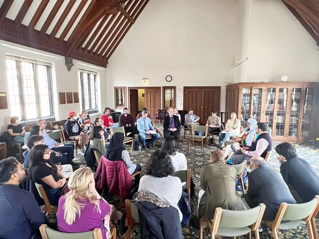 Group of people seated in a circle in a room with high ceilings, wooden beams, and large windows, engaged in a discussion or meeting.