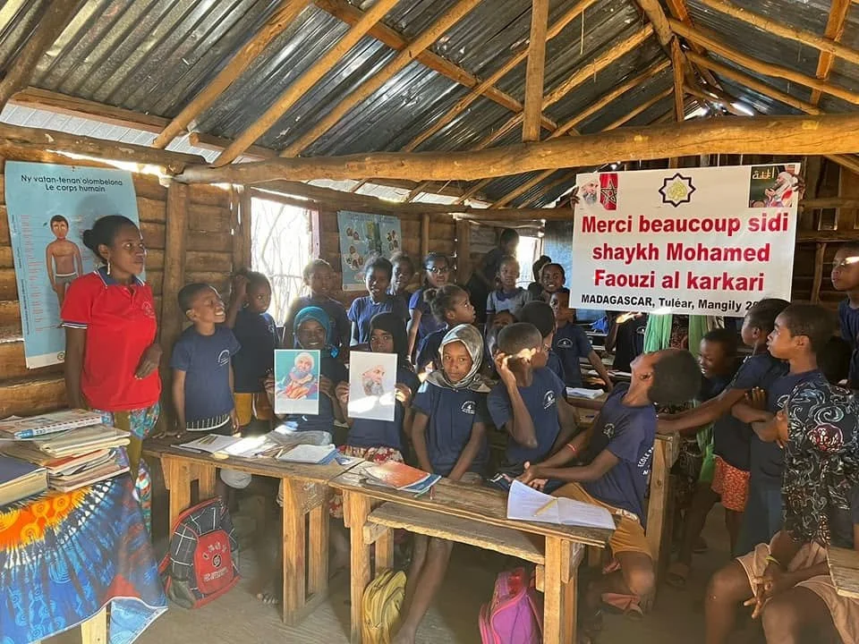 Group of children and adults inside a wooden classroom with educational posters, some children holding drawings, and a large banner thanking Sheikh Mohamed Faouzi al karkari.