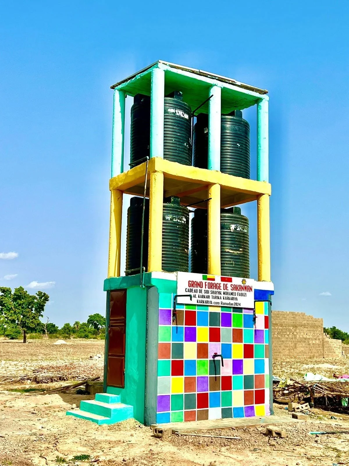 Colorful water distribution structure with black water tanks, painted in vibrant colors, with a sign in French and Arabic indicating a community water project, set against a clear blue sky outside.