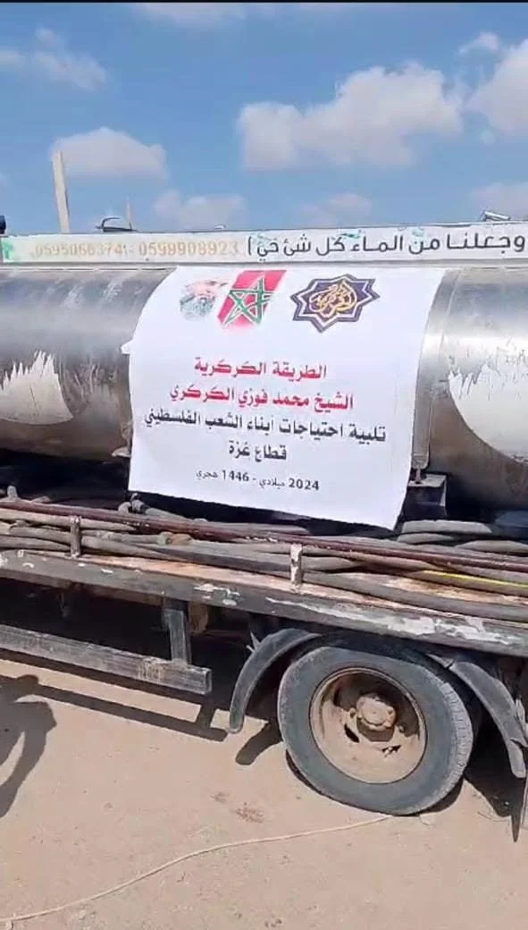 A truck carrying a large banner in Arabic, with the flags of Morocco and Palestine on it, in a desert area with a partly cloudy sky.
