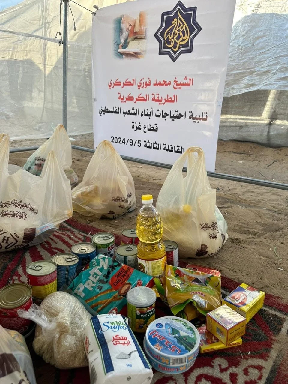 Food and supply donations, including canned goods, packaged snacks, rice, oil, and other food items, placed on a red and black rug outdoors near a sign with Arabic writing.