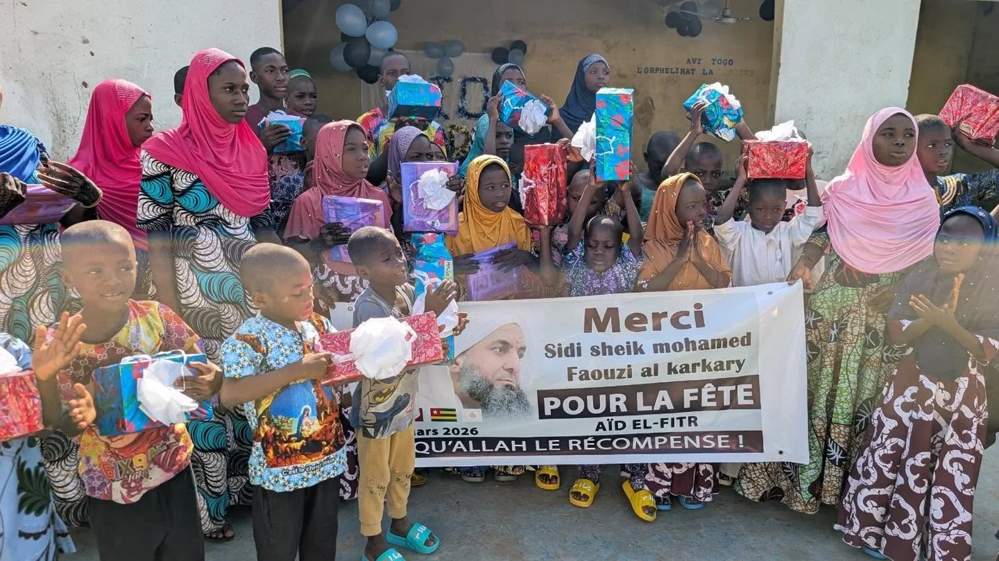 Group of children and women holding gifts and a banner in a celebration; some women are dressed in colorful traditional clothing and headscarves, and children are holding wrapped presents; the banner features a picture of a man and text in French and