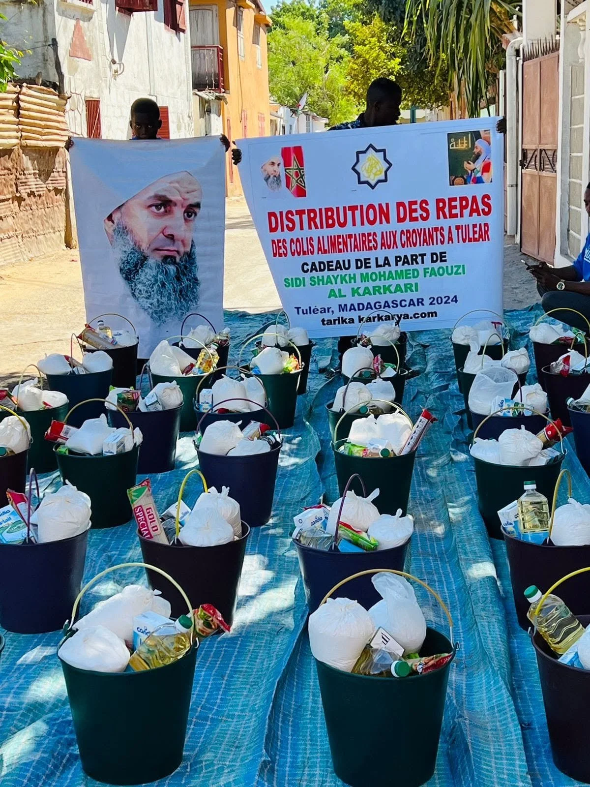 Baskets of food and supplies arranged on a blue tarp on the ground, with a sign in French indicating a meal distribution for believers in Tuléar, Madagascar, 2024.