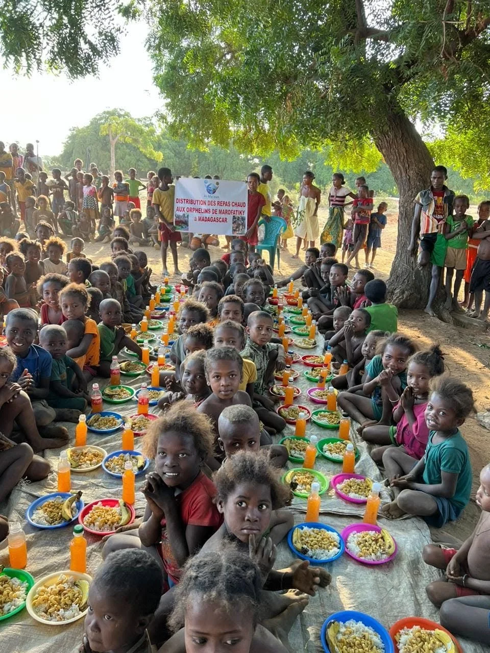 Children sitting on the ground under a large tree, eating a meal with plates and bottles, while others stand in the background holding a banner for a lunch distribution event.